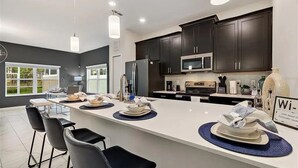 A modern kitchen with dark cabinets, a central island, and a dining area with chairs. - CG236OCA - Luxury 5BR-4BA Villa with Private Pool (Davenport)