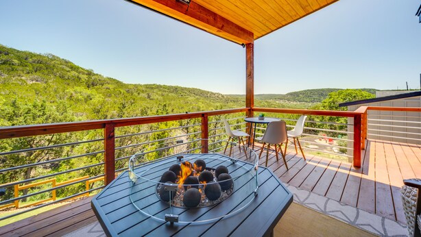 Una terraza de madera con un brasero, mobiliario exterior y una vista panorámica de una ladera boscosa.