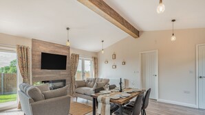 A modern living room with a fireplace, a dining area with a wooden table and chairs, and a large window with curtains. - West End Farm Lodge Alabaster (Ruardean, Forest of Dean)