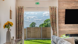 A room with a wooden floor, a sofa, a TV, a vase with flowers, and a view of the outdoors. - West End Farm Lodge Alabaster (Ruardean, Forest of Dean)