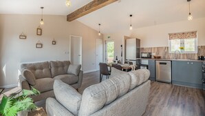 A spacious living area with a sofa set, a dining table with chairs, a kitchen area with a microwave, and a window with a view of greenery. - West End Farm Lodge Alabaster (Ruardean, Forest of Dean)