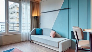 A modern hotel room with a sofa, a chair, a table, a lamp, and a view of a building through a window.