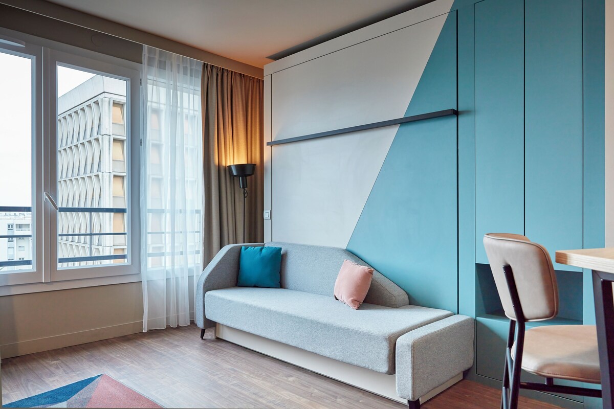 A modern hotel room with a sofa, a chair, a table, a lamp, and a view of a building through a window.