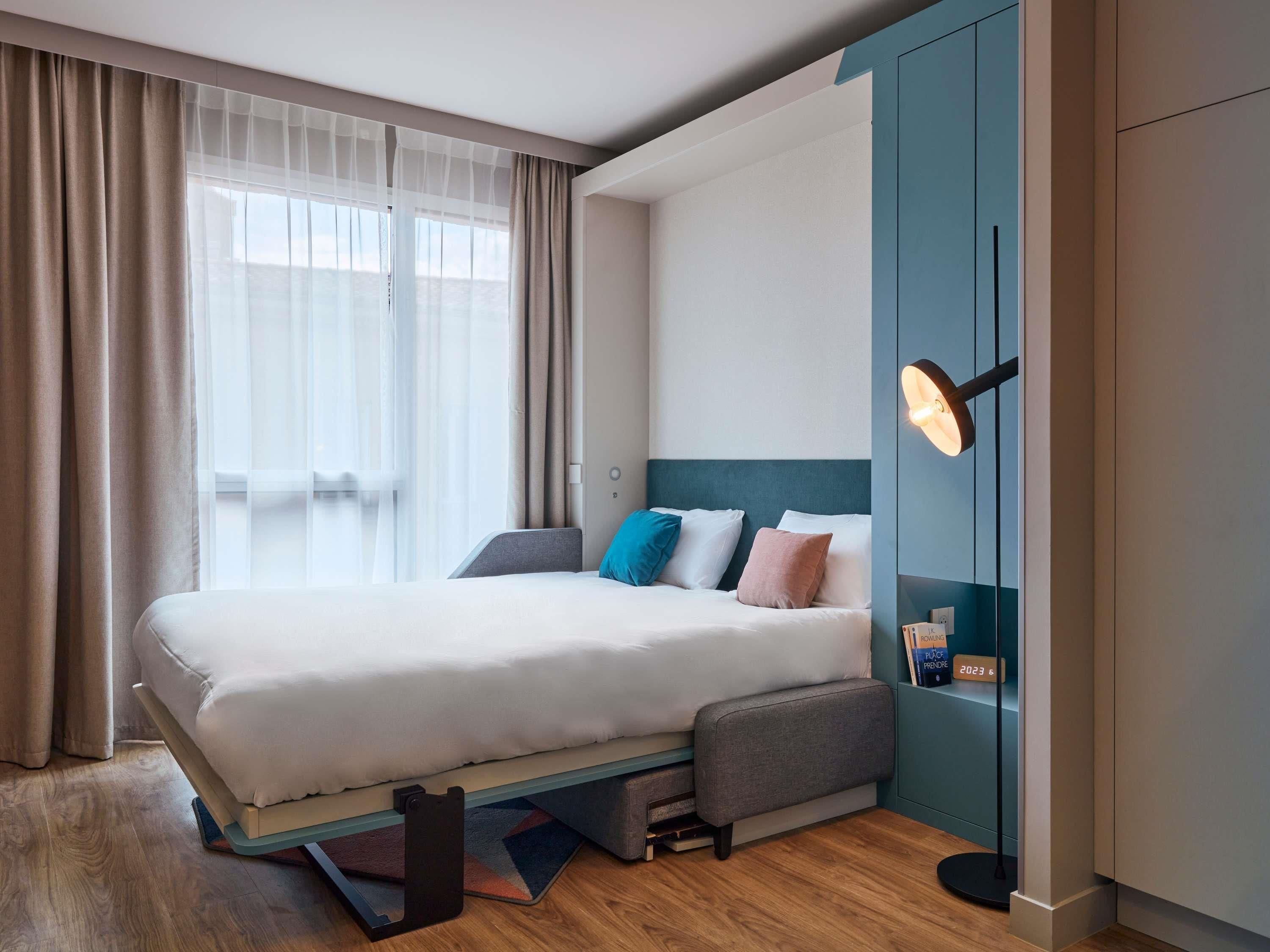 A modern hotel room with a large bed, a bedside table, a nightstand, a wall-mounted reading light, and a view of the sea through sheer curtains.