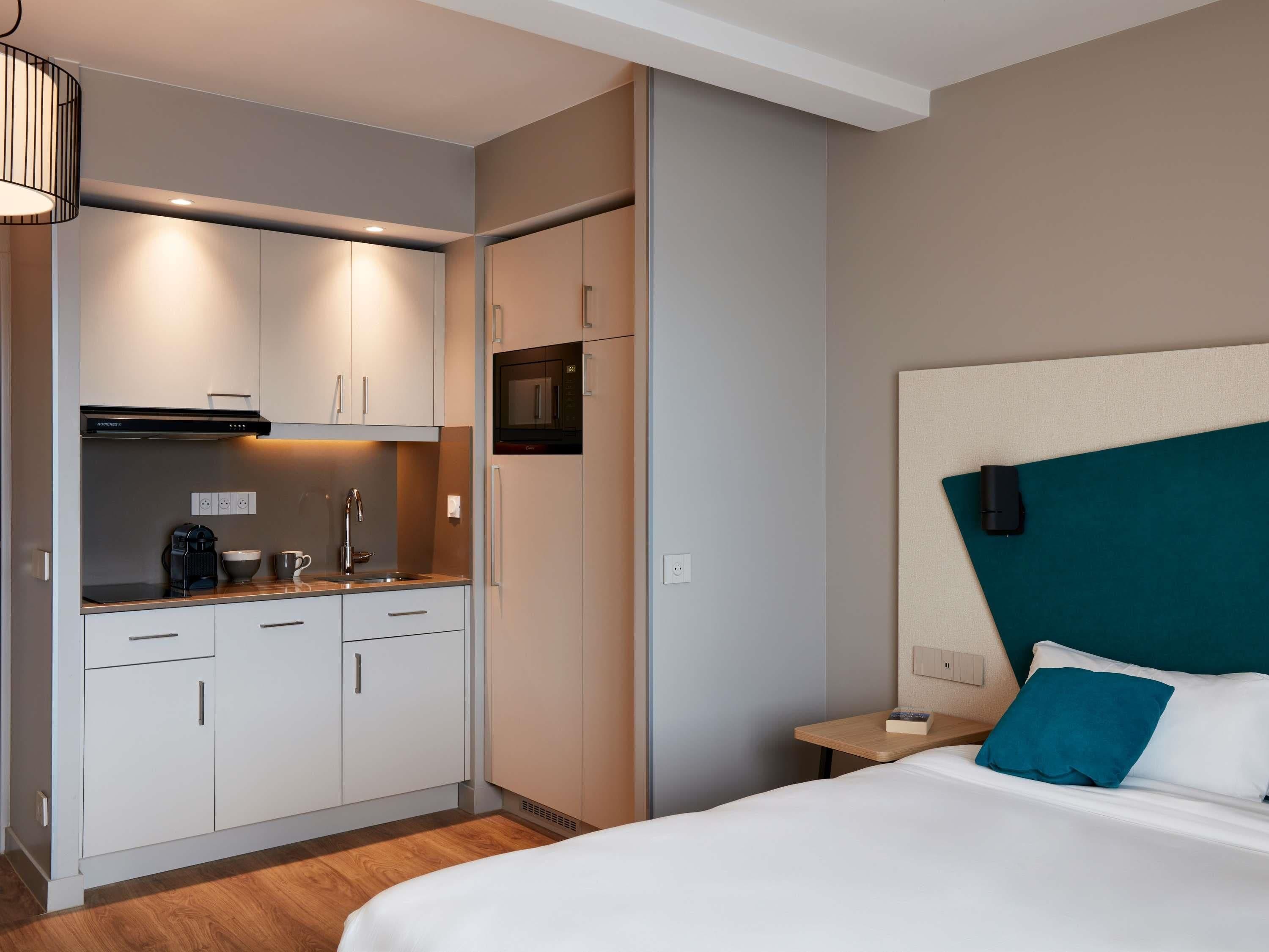 A hotel room with a kitchenette, a bed with teal and white pillows, and a wooden nightstand.