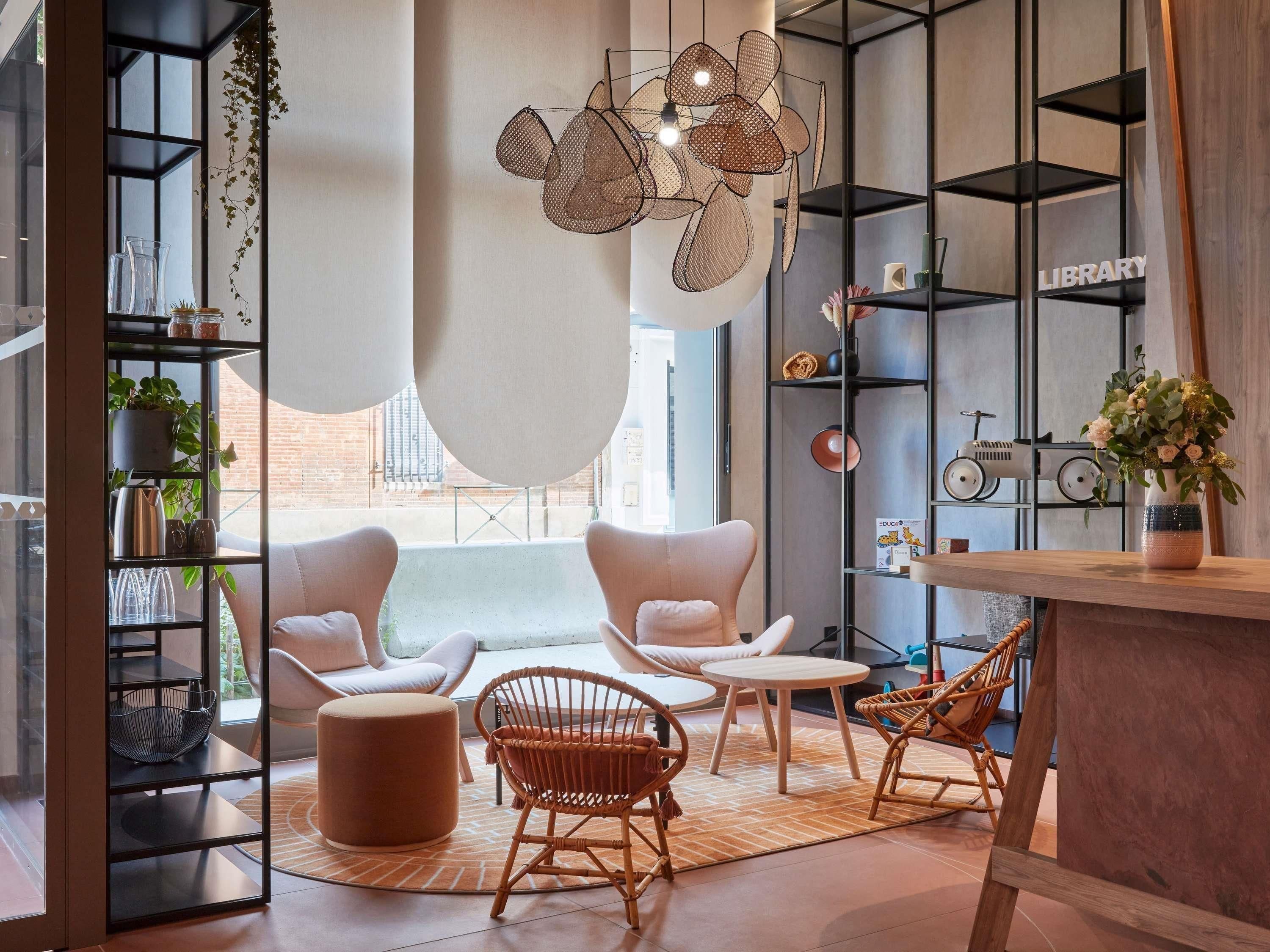 A modern living room with a sofa set, a coffee table, and a dining area with chairs. There are shelves with books and decorative items, and a large window providing natural light.