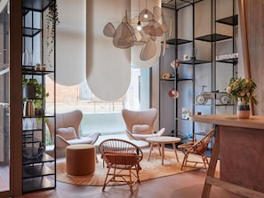 A modern living room with a sofa set, a coffee table, and a dining area with chairs. There are shelves with books and decorative items, and a large window providing natural light.