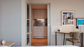 Habitación de hotel con cama, escritorio con silla, mesita con reloj, kitchenette y un cuadro en la pared.