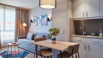 A modern living room with a sofa, a dining table with chairs, a kitchen area with cabinets, and a large window with curtains.