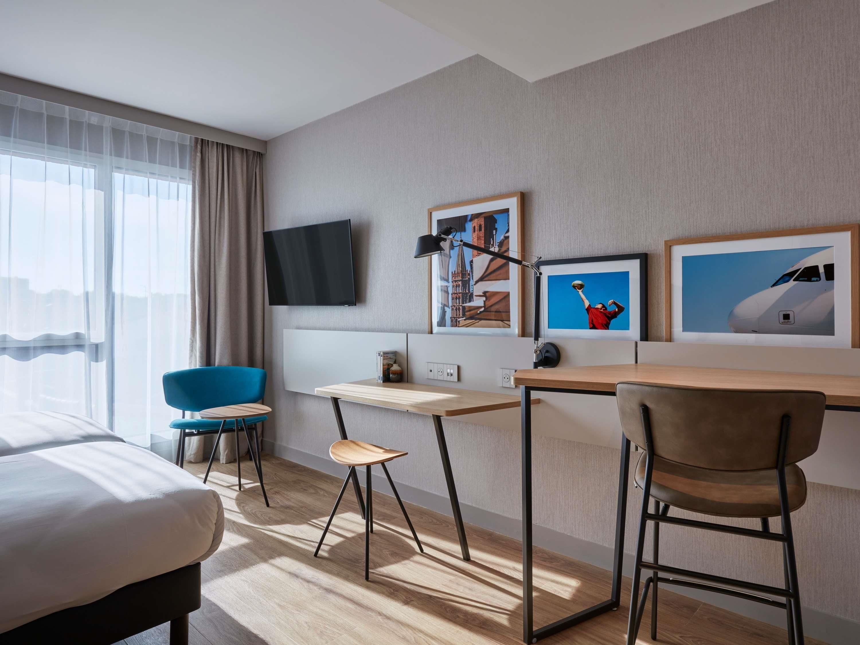 A hotel room with a bed, a desk, a chair, a TV, and artwork on the wall.