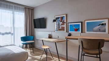 A hotel room with a bed, a desk, a chair, a TV, and artwork on the wall.