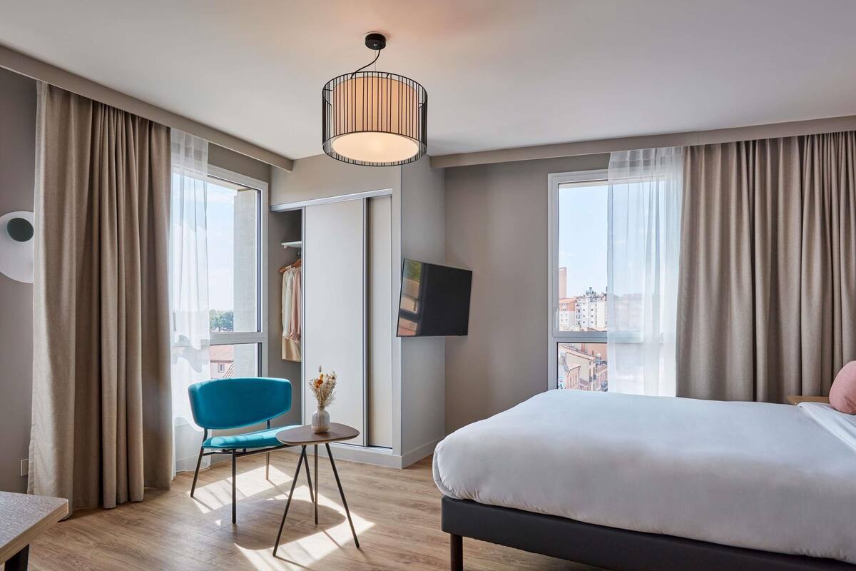 A modern hotel room with a large bed, a blue chair, a small table, and a TV on the wall.