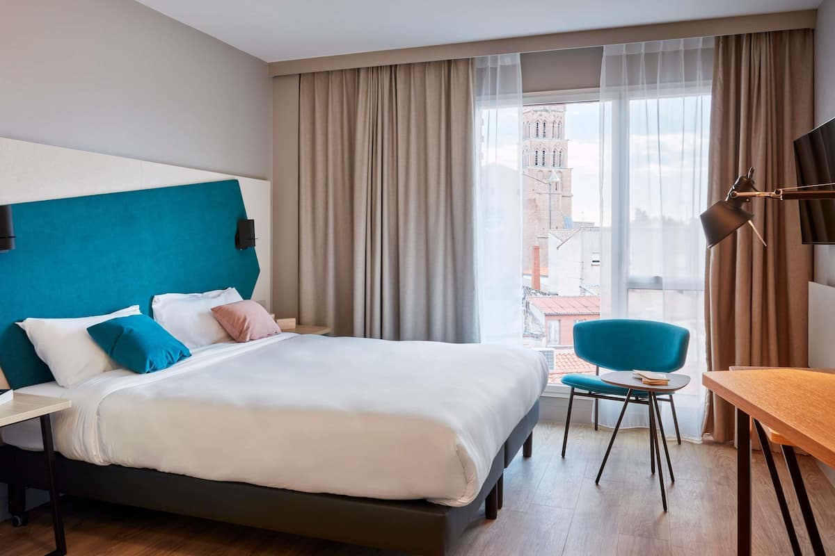 Una camera d'albergo con un letto, una scrivania, una sedia e vista sugli edifici dalla finestra.