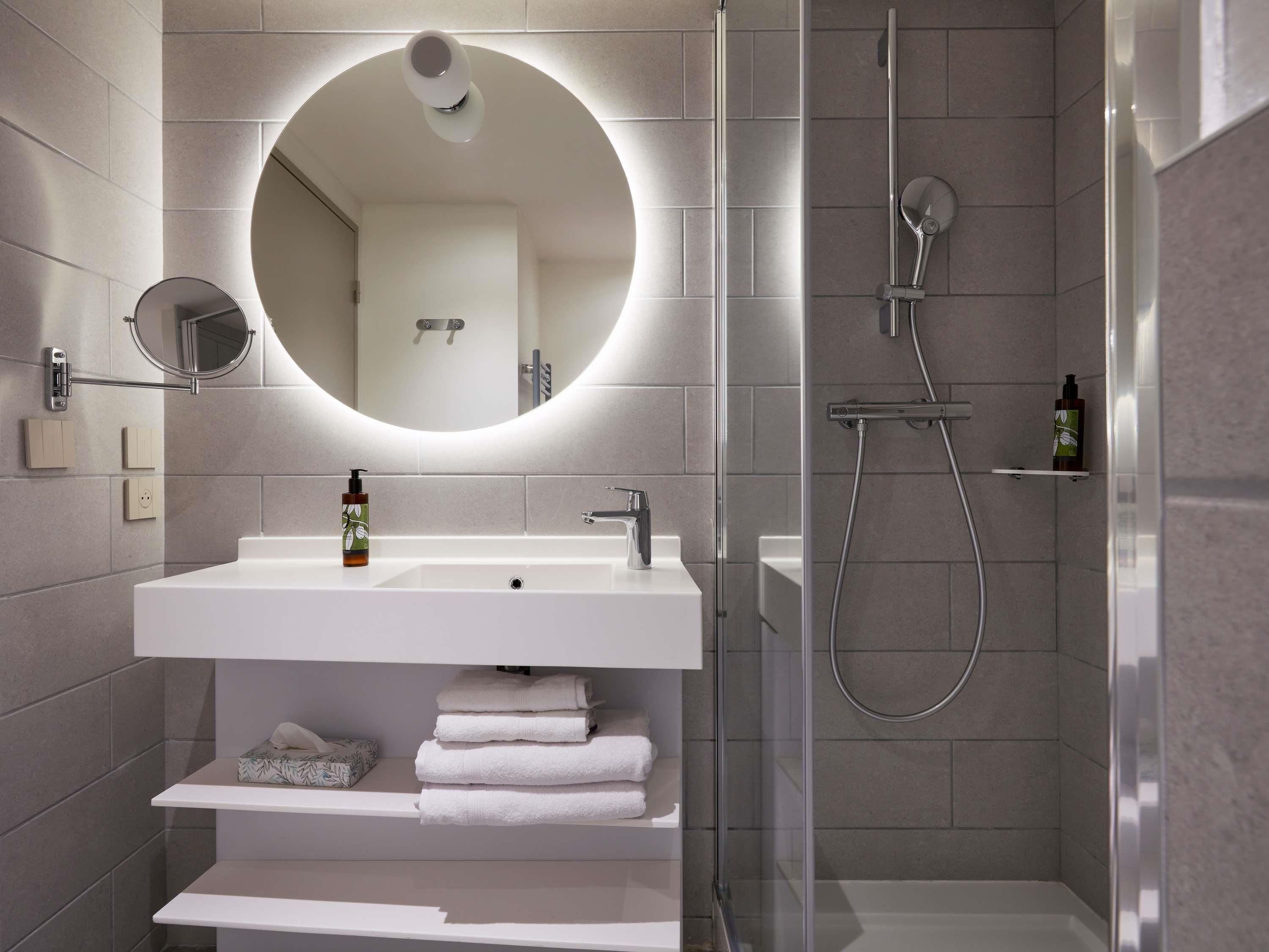 Un bagno moderno con un lavandino bianco, uno specchio rotondo, un soffione doccia estraibile e una cabina doccia in vetro.