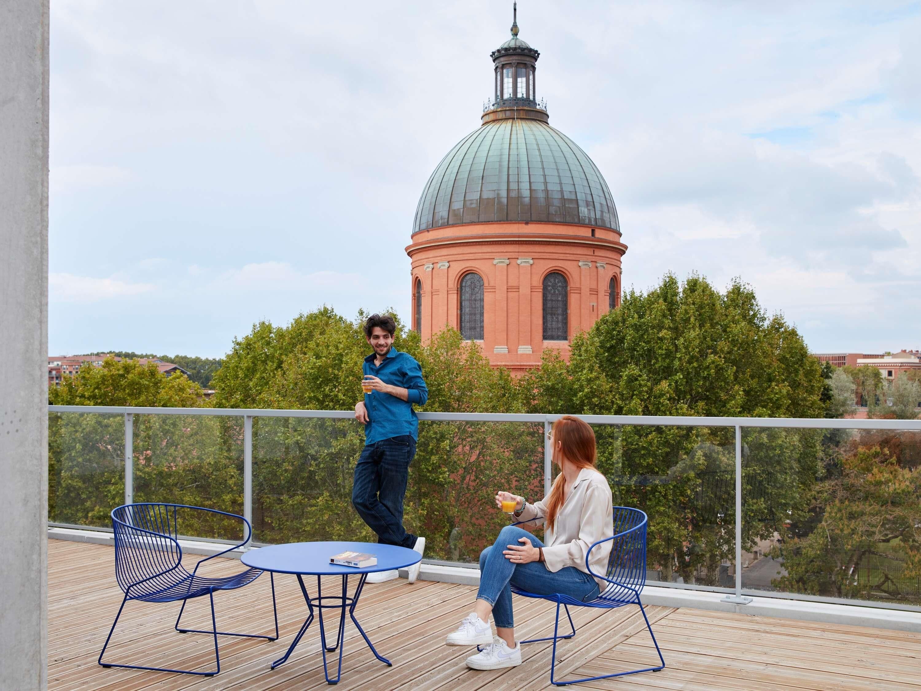 En balkon med to blå stole og et lille bord, med udsigt til en historisk bygning med en kuppel og buede vinduer.