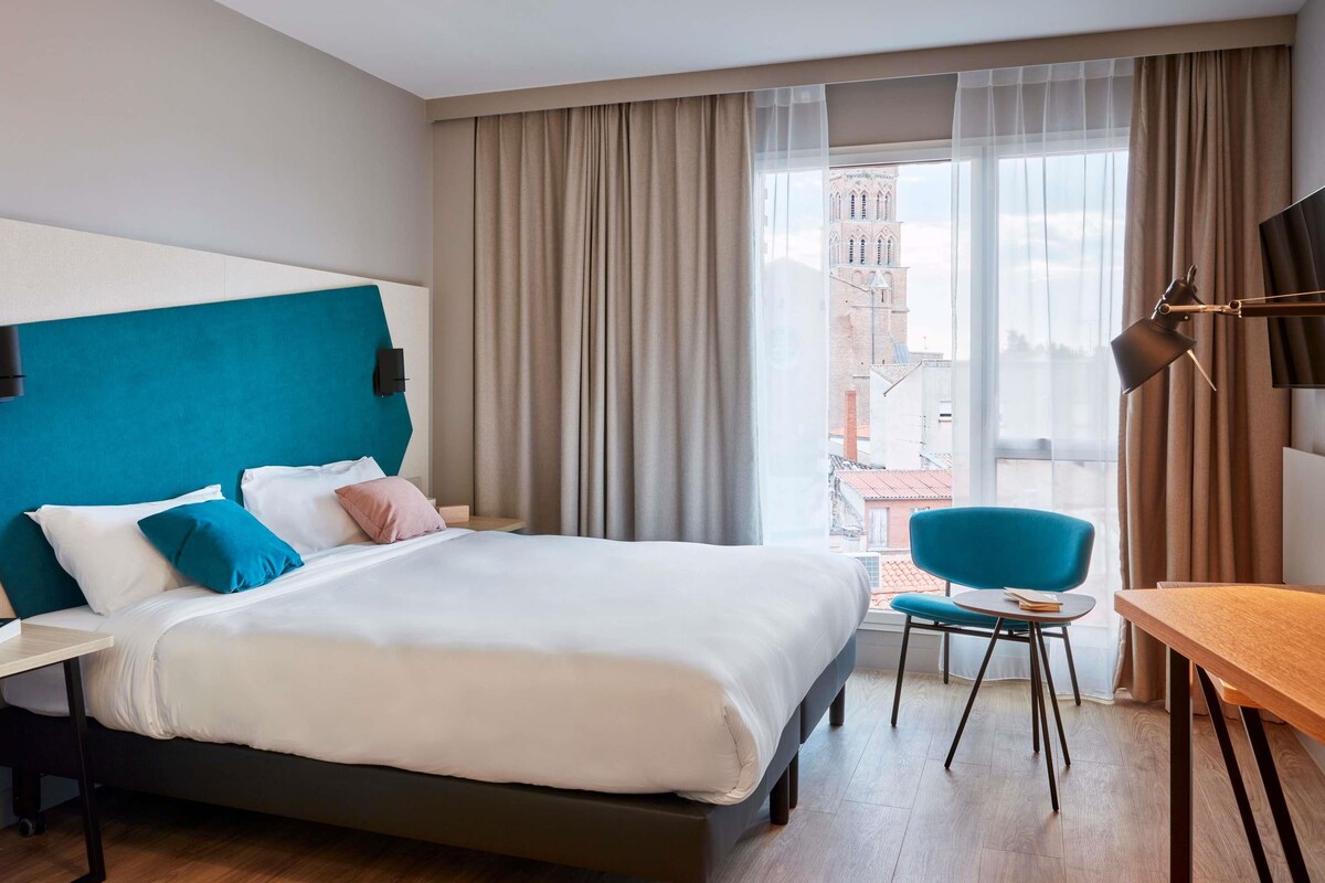 Une chambre d’hôtel avec un lit, un bureau, une chaise et une vue sur les immeubles par la fenêtre.