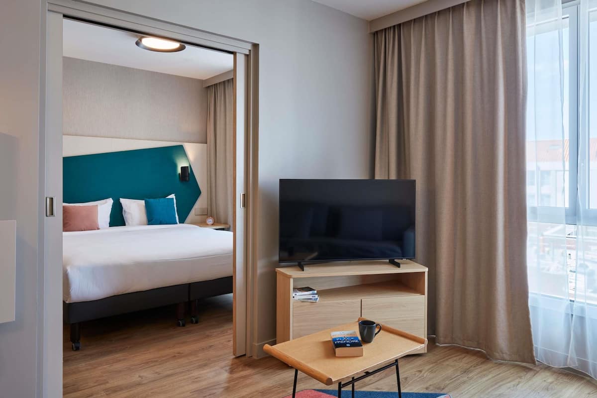 Una camera d'albergo con un letto, una televisione su un mobiletto in legno, un tavolino con un vassoio e una finestra con tende.