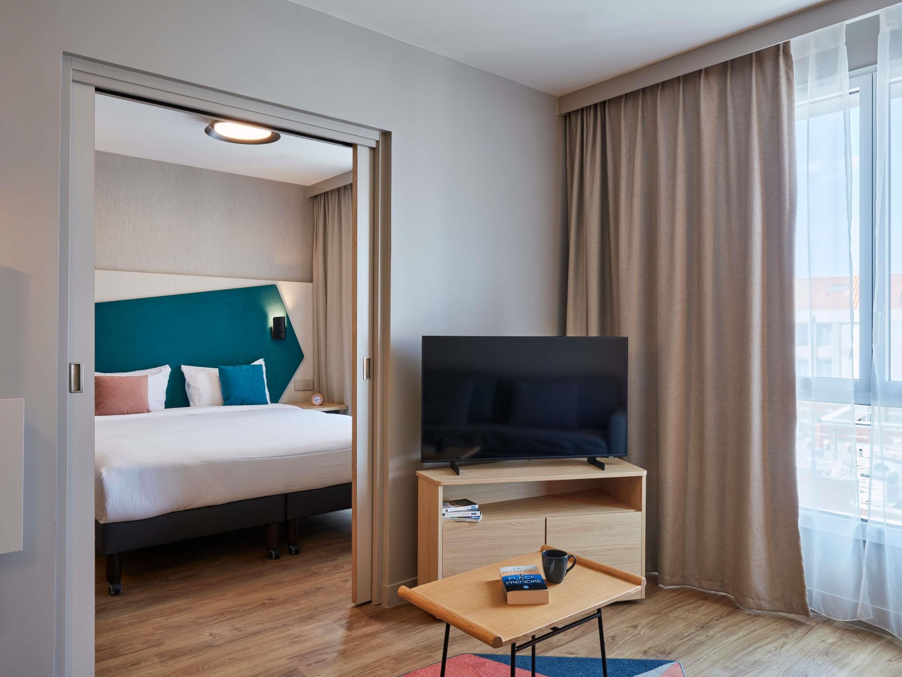 A hotel room with a bed, a television on a wooden stand, a small table with a tray, and a window with curtains.