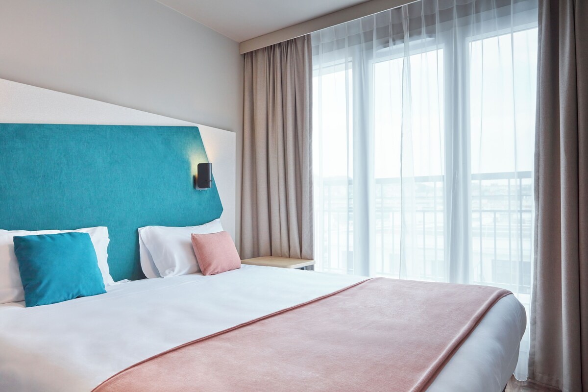 A hotel room with a large bed, a headboard, and a view of the city through sheer curtains.