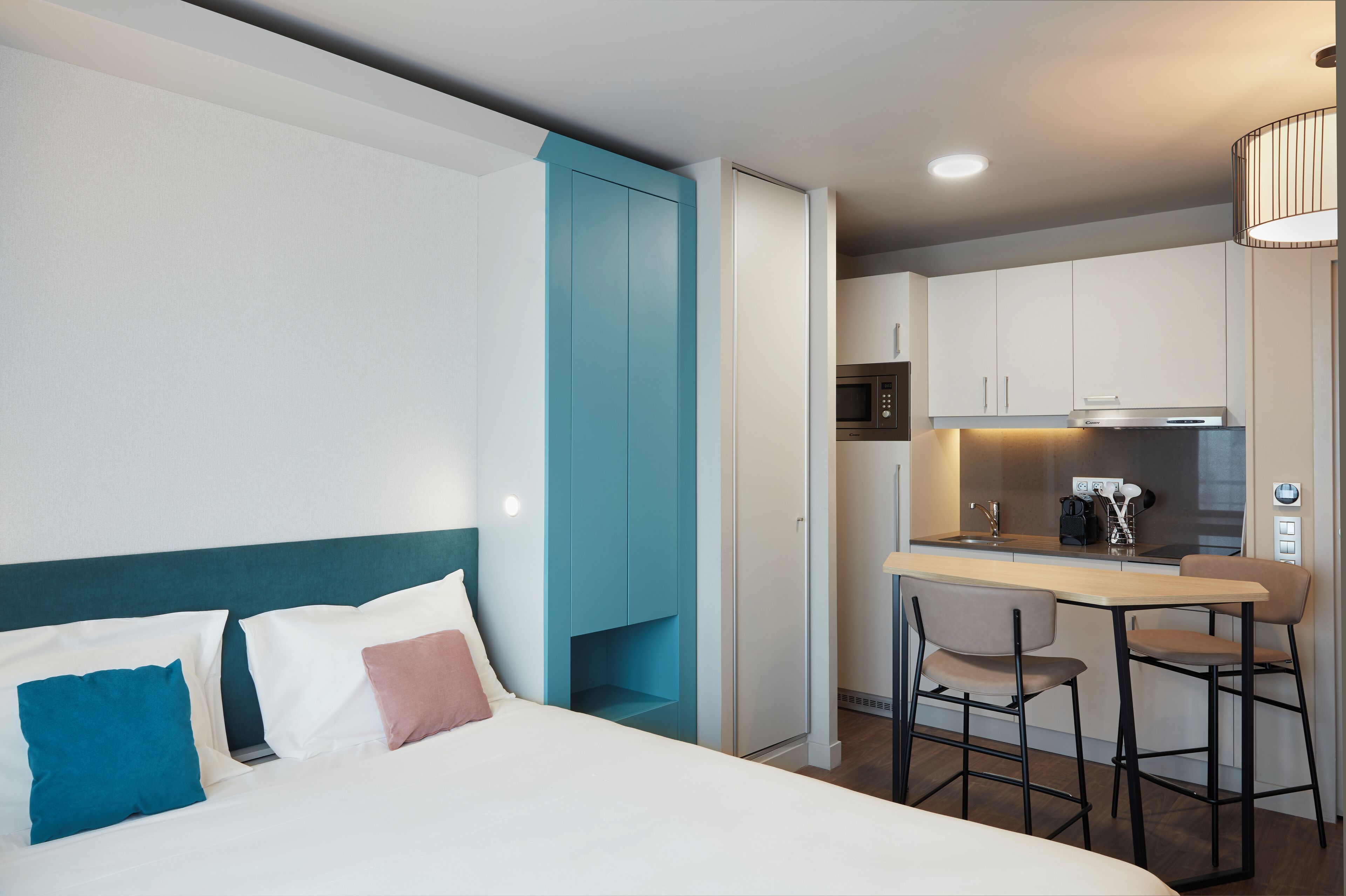 A modern hotel room with a bed, a small kitchenette, and a dining area.