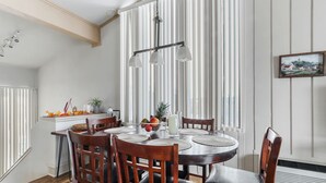 A dining area with a round table, wooden chairs, a chandelier, and a window with vertical blinds.
