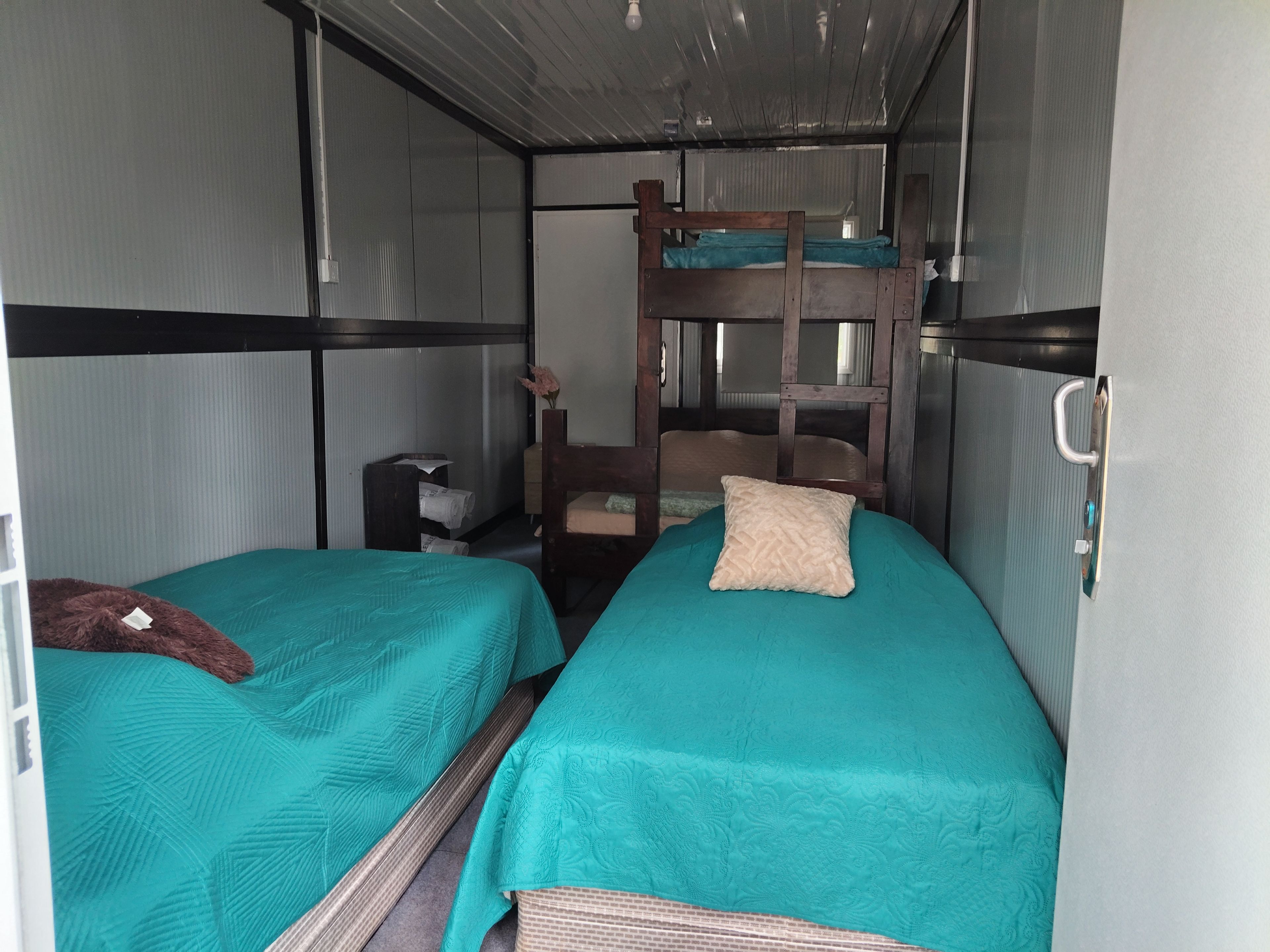 Habitación pequeña con dos camas, una litera, una mesita y un estante en la pared.