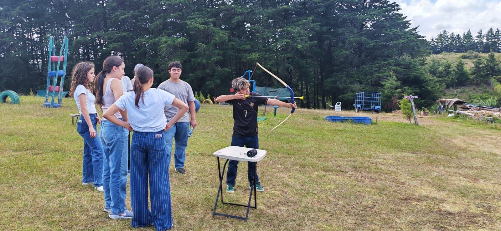 Escena al aire libre con una persona apuntando un arco a una diana, rodeada de un grupo de personas.