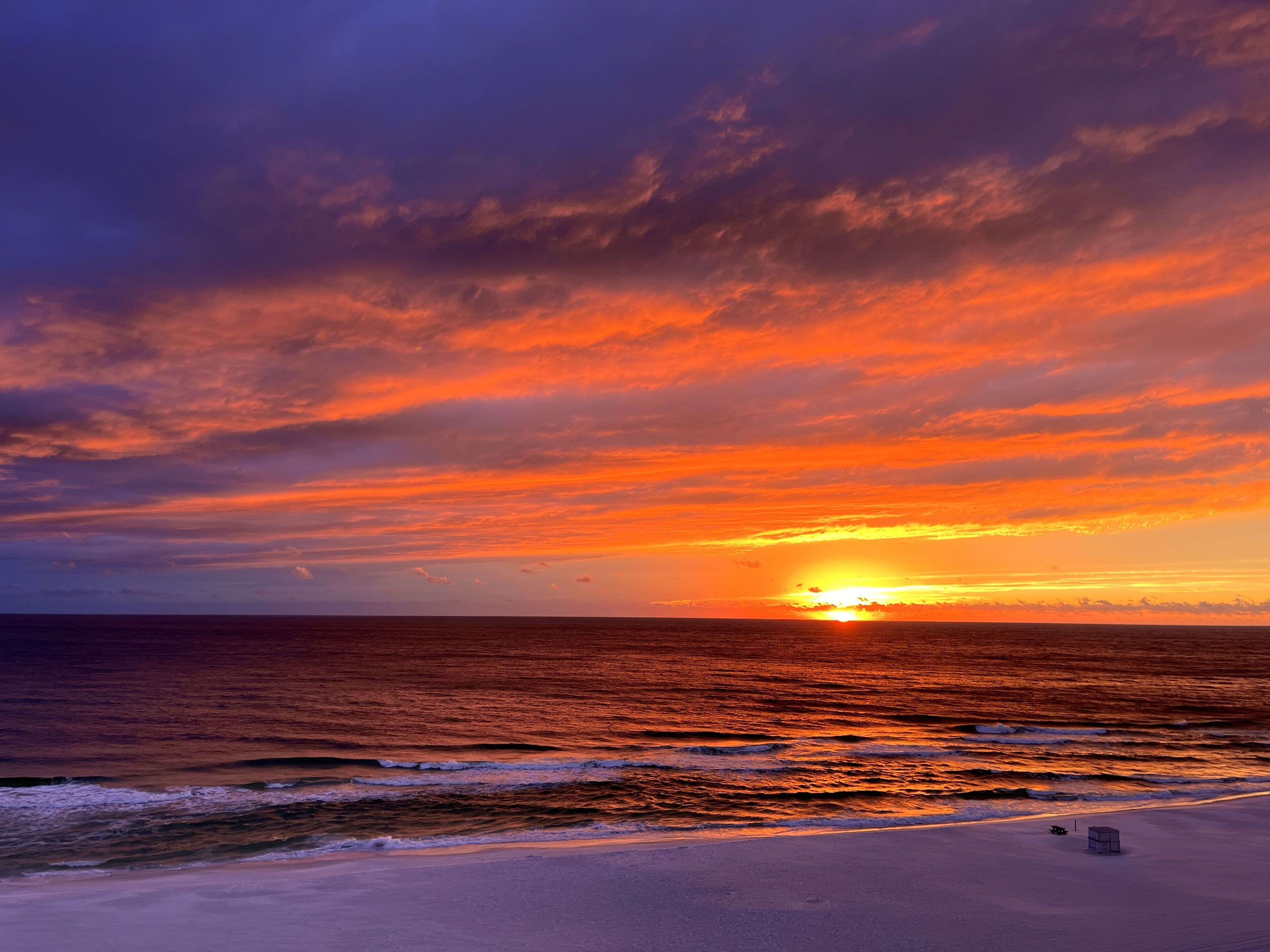 A beach with a sunset over the ocean, a clear sky, and a small structure on the sand.