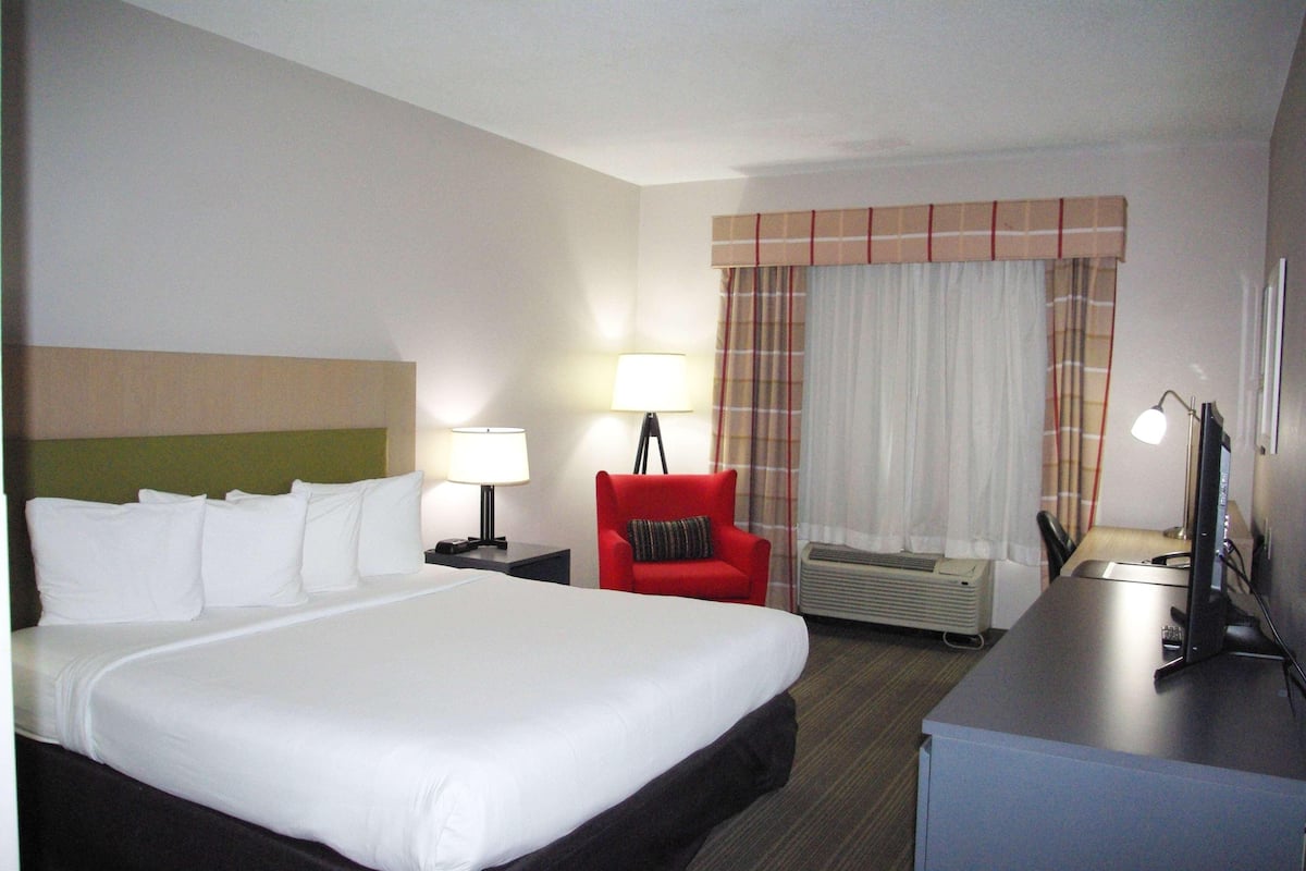 A hotel room with a large bed, a desk with a computer, a chair, a lamp, and an air conditioning unit.