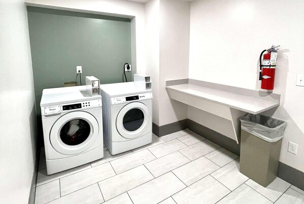 A laundry area with two front-loading washing machines, a grey trash bin, a white sink, and a red fire extinguisher mounted on the wall.