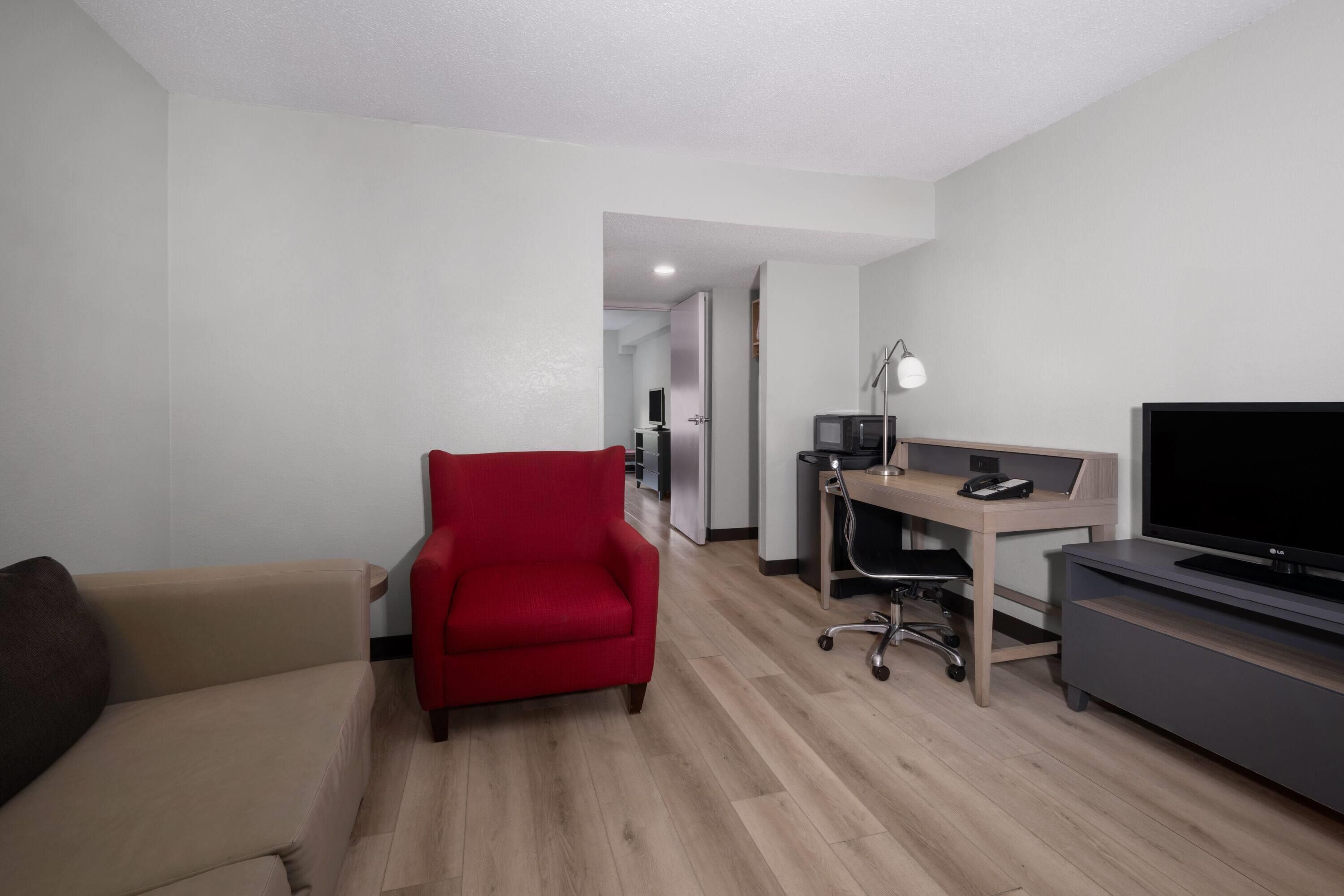 A hotel room with a sofa, a red armchair, a desk with a computer, a TV, and a lamp.