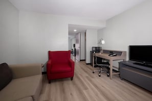 Habitación de hotel con un sofá, un sillón rojo, un escritorio con computadora, un televisor y una lámpara.