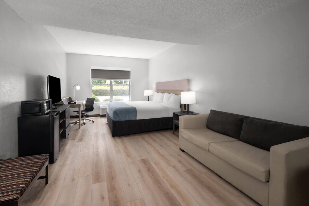 A modern hotel room with a bed, a sofa, a desk with a computer, a TV, and a window with a view.