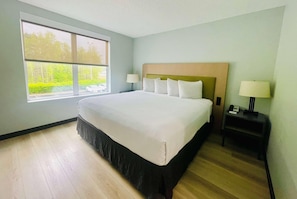 Une chambre dâhĂŽtel avec un grand lit, une fenĂȘtre donnant sur un paysage verdoyant et une table de chevet avec une lampe.