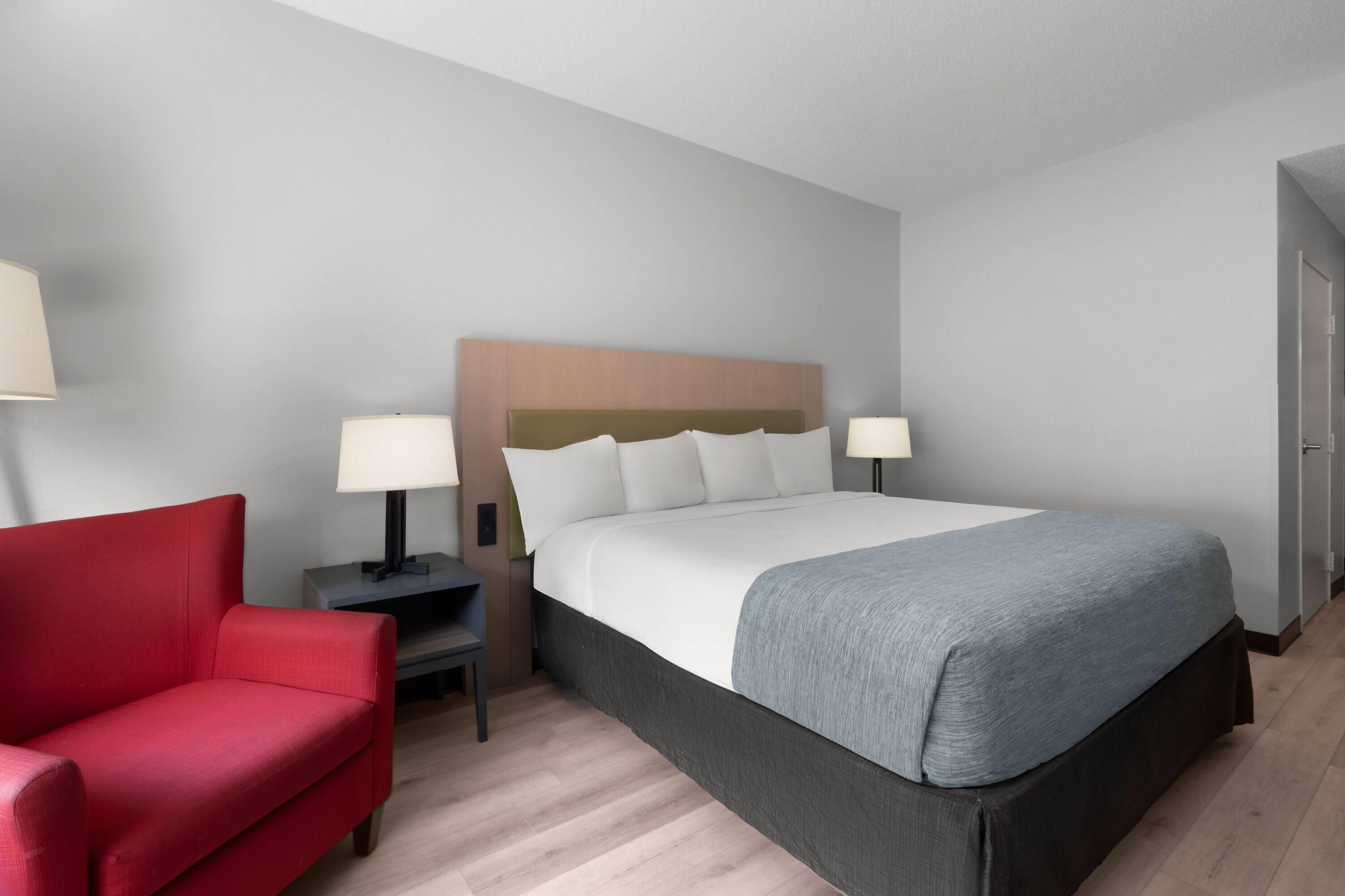 Une chambre d’hôtel avec un lit, une chaise rouge, deux tables de chevet avec des lampes et un plancher en bois.