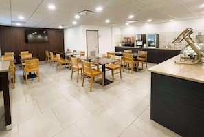 A restaurant with wooden tables and chairs, a TV mounted on the wall, and a counter with a coffee machine. - Wingate By Wyndham Matthews Nc (Matthews)