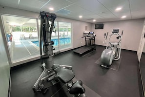 Une salle de sport Ă©quipĂ©e de matĂ©riel dâexercice, dâun tapis de course et offrant une vue sur une piscine extĂ©rieure.
