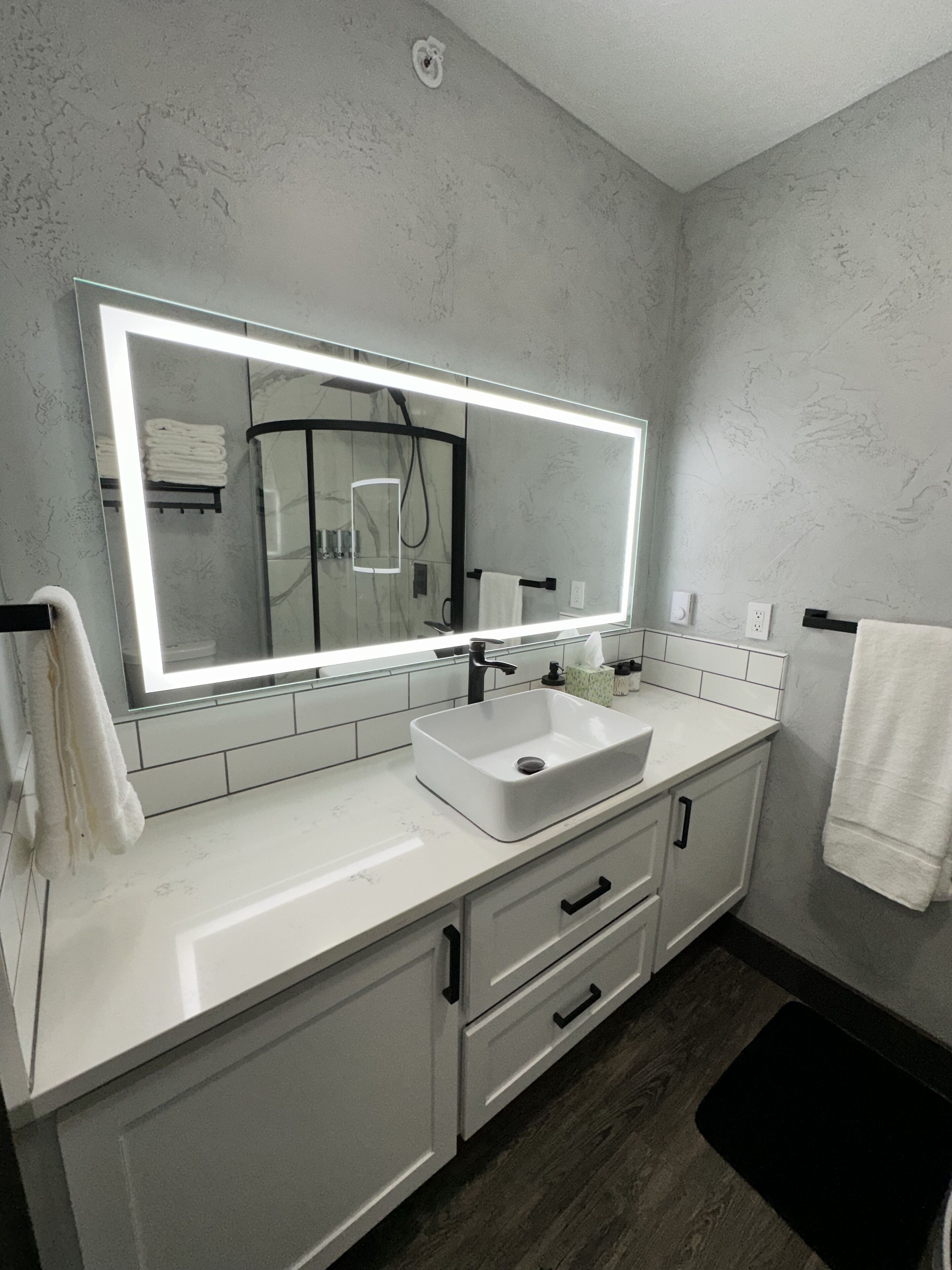 Une salle de bain moderne avec un grand miroir, un lavabo blanc et un revêtement de sol en bois foncé.
