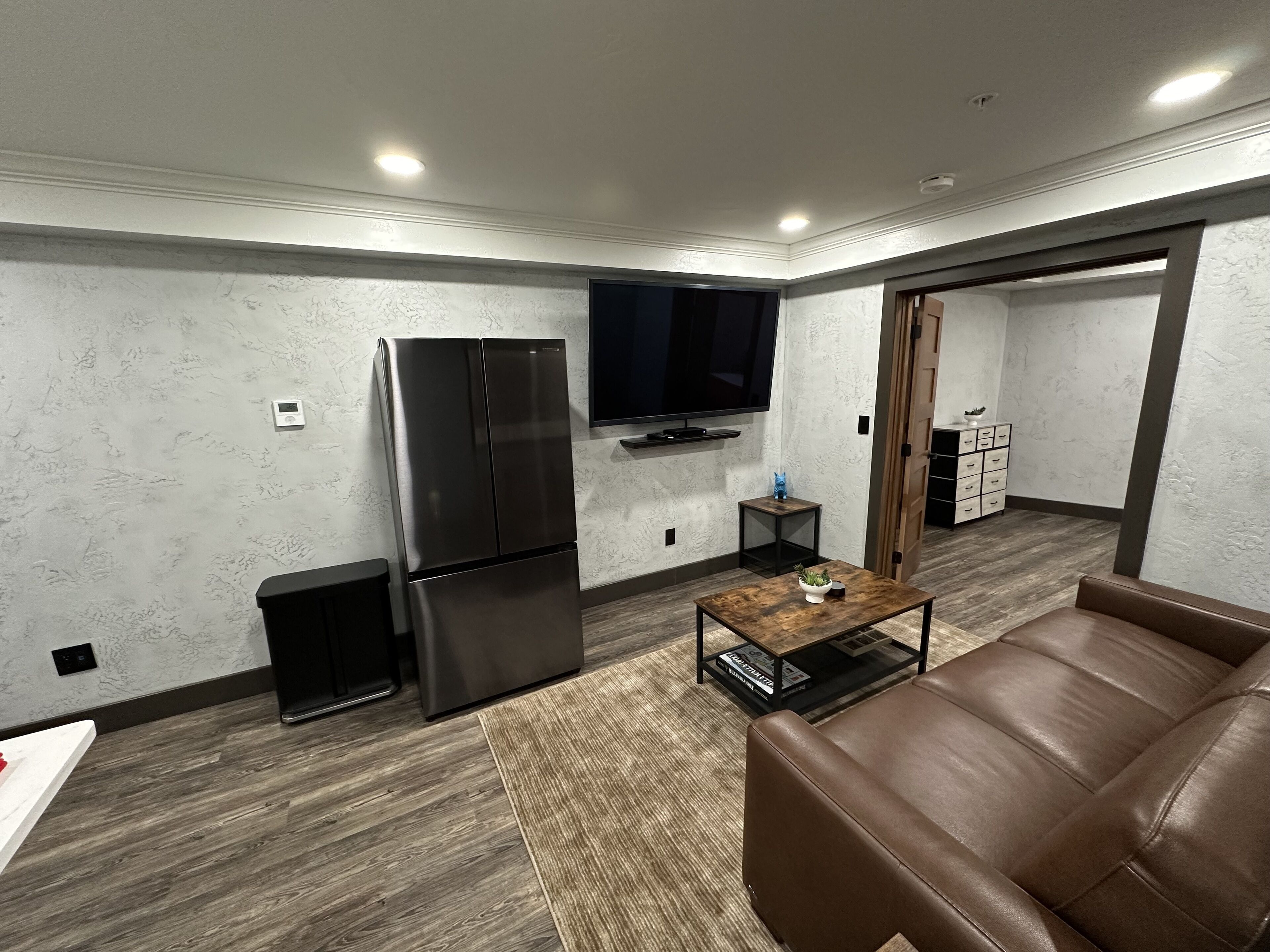 A modern living room with a large flat-screen TV, a stainless steel refrigerator, a brown leather sofa, and a small coffee table.