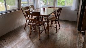 A dining area with a wooden table and chairs, large windows with white curtains, and a view of trees and houses outside. - Webster Camp - rustic getaway with Webster Lake access, fire pit & large deck!   (Franklin)