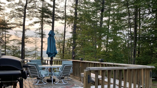 A wooden deck with a table and chairs, surrounded by tall trees. - Webster Camp - rustic getaway with Webster Lake access, fire pit & large deck!   (Franklin)