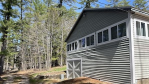 A single-story building with a garage door, surrounded by a forested area. - Webster Camp - rustic getaway with Webster Lake access, fire pit & large deck!   (Franklin)