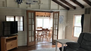 A spacious living room with a high ceiling, wooden beams, a television on a wooden cabinet, a grey sofa, a white coffee table, and a patterned rug. - Webster Camp - rustic getaway with Webster Lake access, fire pit & large deck!   (Franklin)