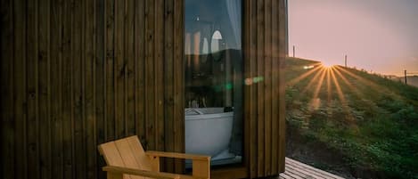 Una cabaña de madera con baño de cristal, una terraza de madera y vistas a un atardecer sobre una colina cubierta de hierba.