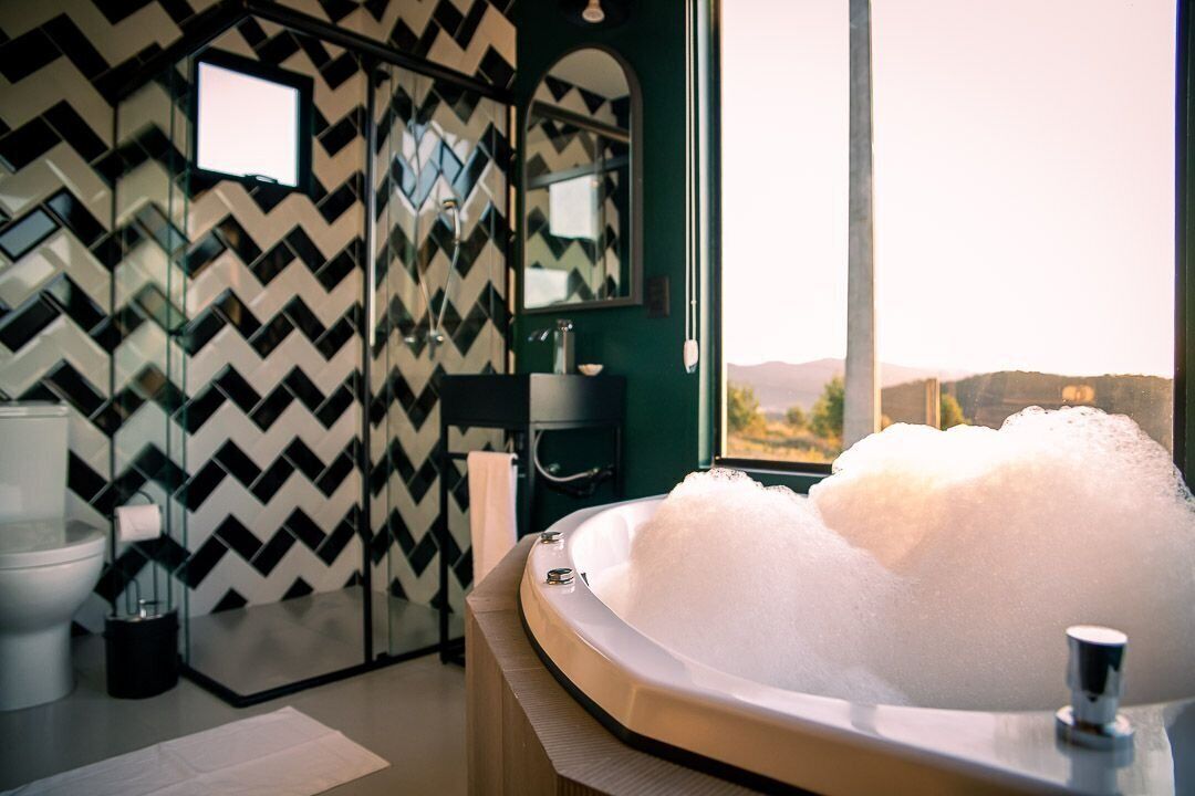 A modern bathroom with a large bathtub filled with bubbles, a mirror, and a view of the outdoors.