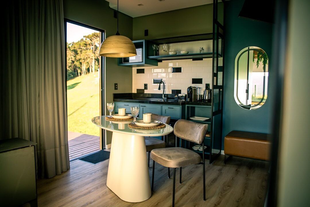 A modern kitchen with a dining area, featuring a round table with chairs, a hanging light fixture, and a view of the outdoors.