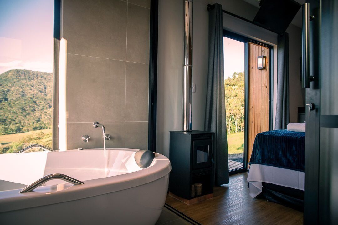 A modern bathroom with a large bathtub, a wooden door leading outside, and a bed with a blue bedspread.