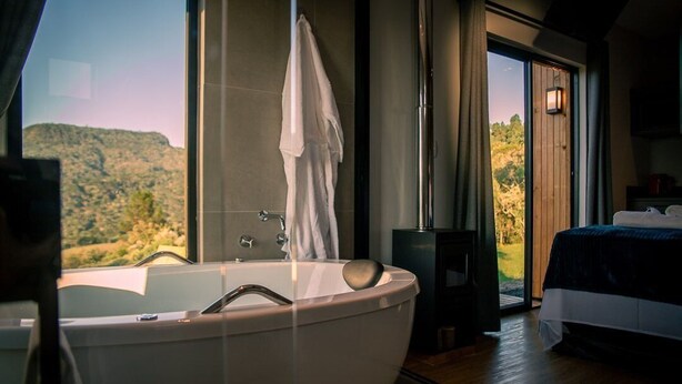 A modern bathroom with a freestanding bathtub, a large window offering a view of a mountainous landscape, and a separate bedroom with a bed and a nightstand. - Cabanas Sunset View (Urubici)