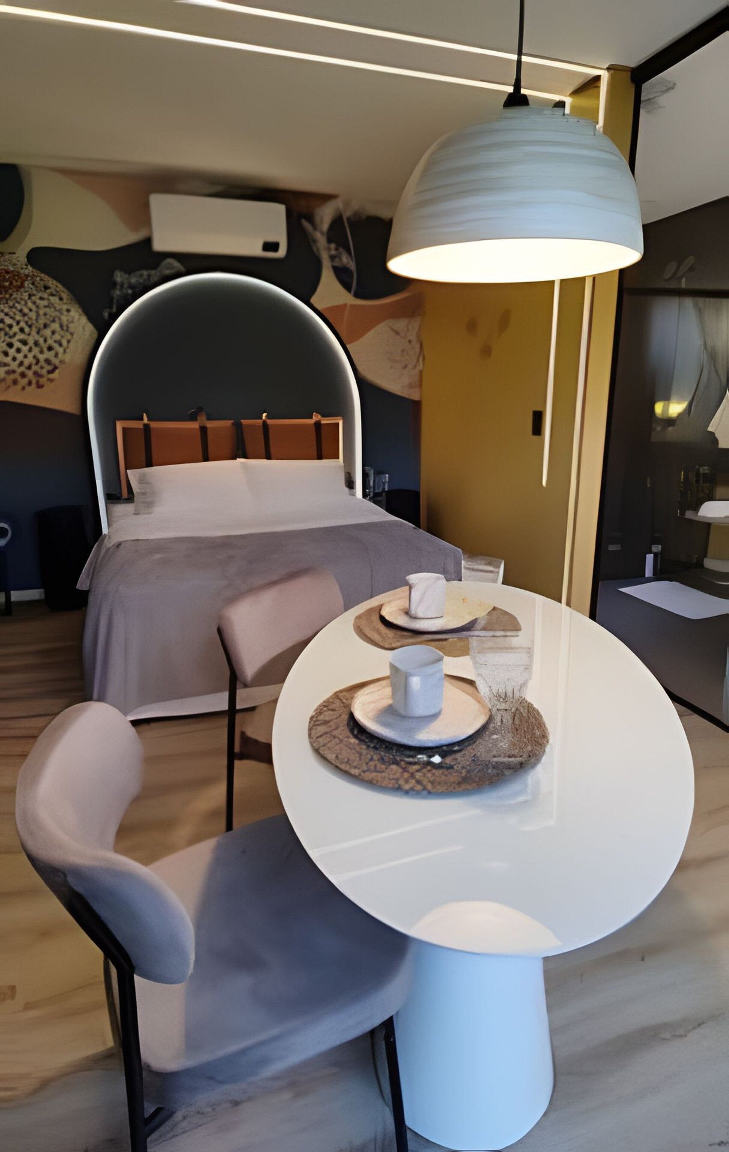A modern hotel room with a bed, a dining table with chairs, and a hanging light fixture.