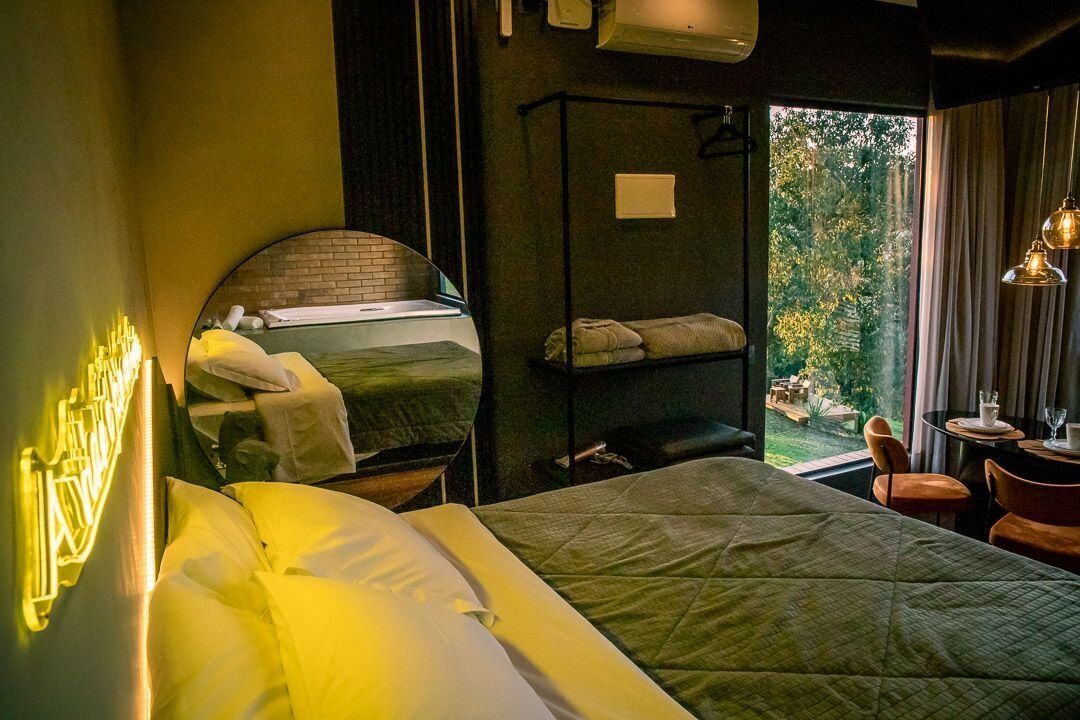 Una camera d'albergo con un letto, un comodino, uno specchio, una finestra vista verde e un tavolino con una sedia.
